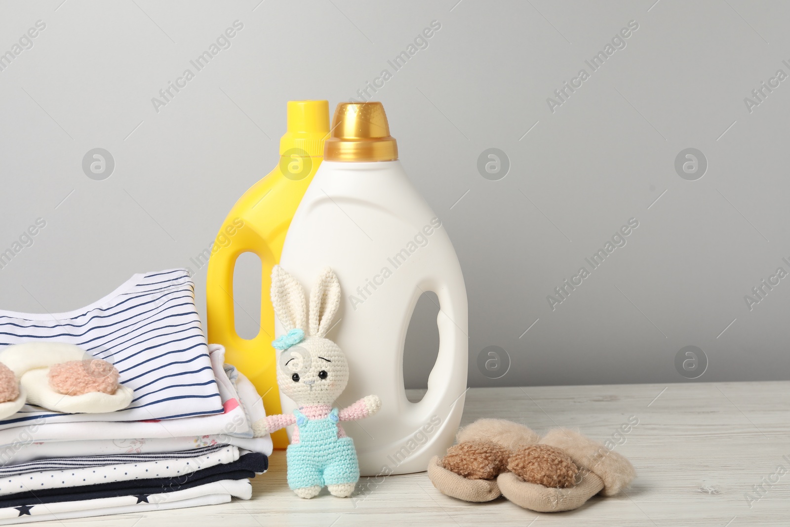 Bottles of fabric softeners, baby clothes and toy on light wooden table against grey background Photo of Bottles of fabric softeners, baby clothes and toy on light wooden table against grey background