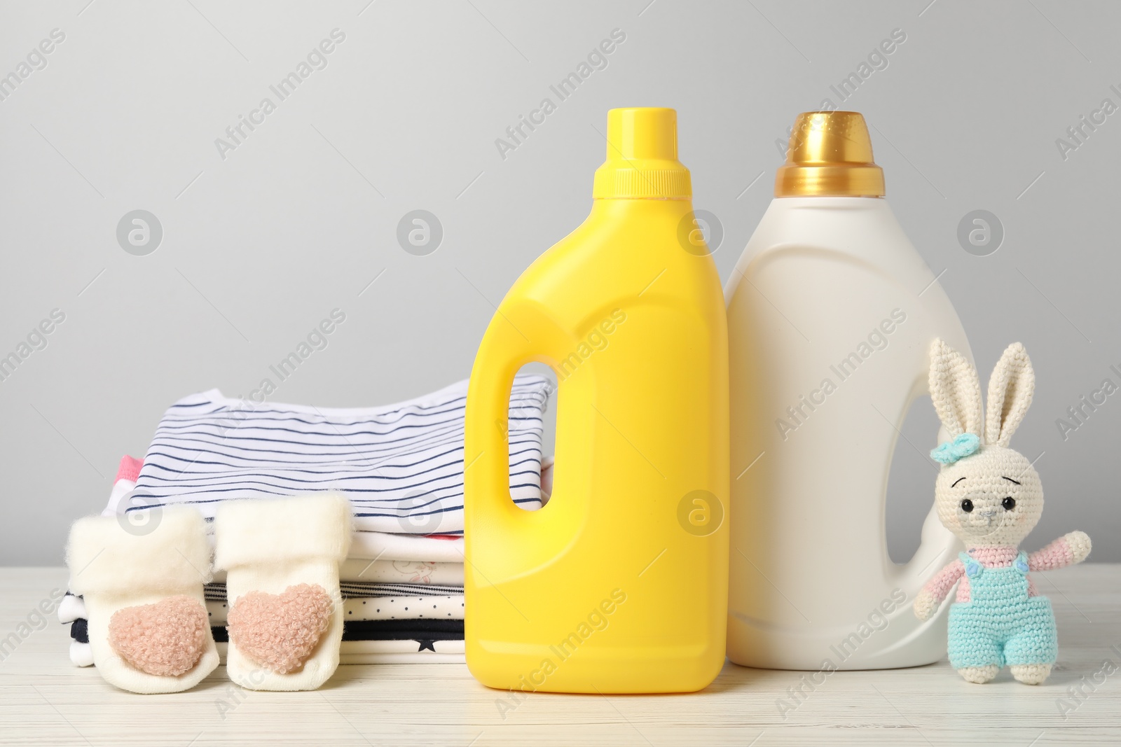 Bottles of fabric softeners, baby clothes and toy on light wooden table against grey background Photo of Bottles of fabric softeners, baby clothes and toy on light wooden table against grey background