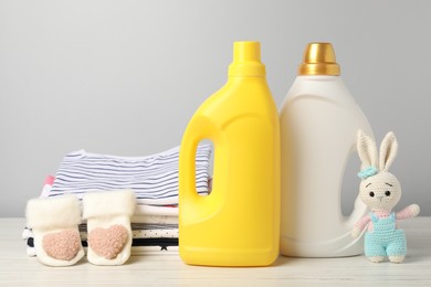 Photo of Bottles of fabric softeners, baby clothes and toy on light wooden table against grey background