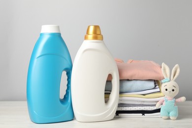 Photo of Bottles of fabric softeners, baby clothes and toy on light wooden table against grey background