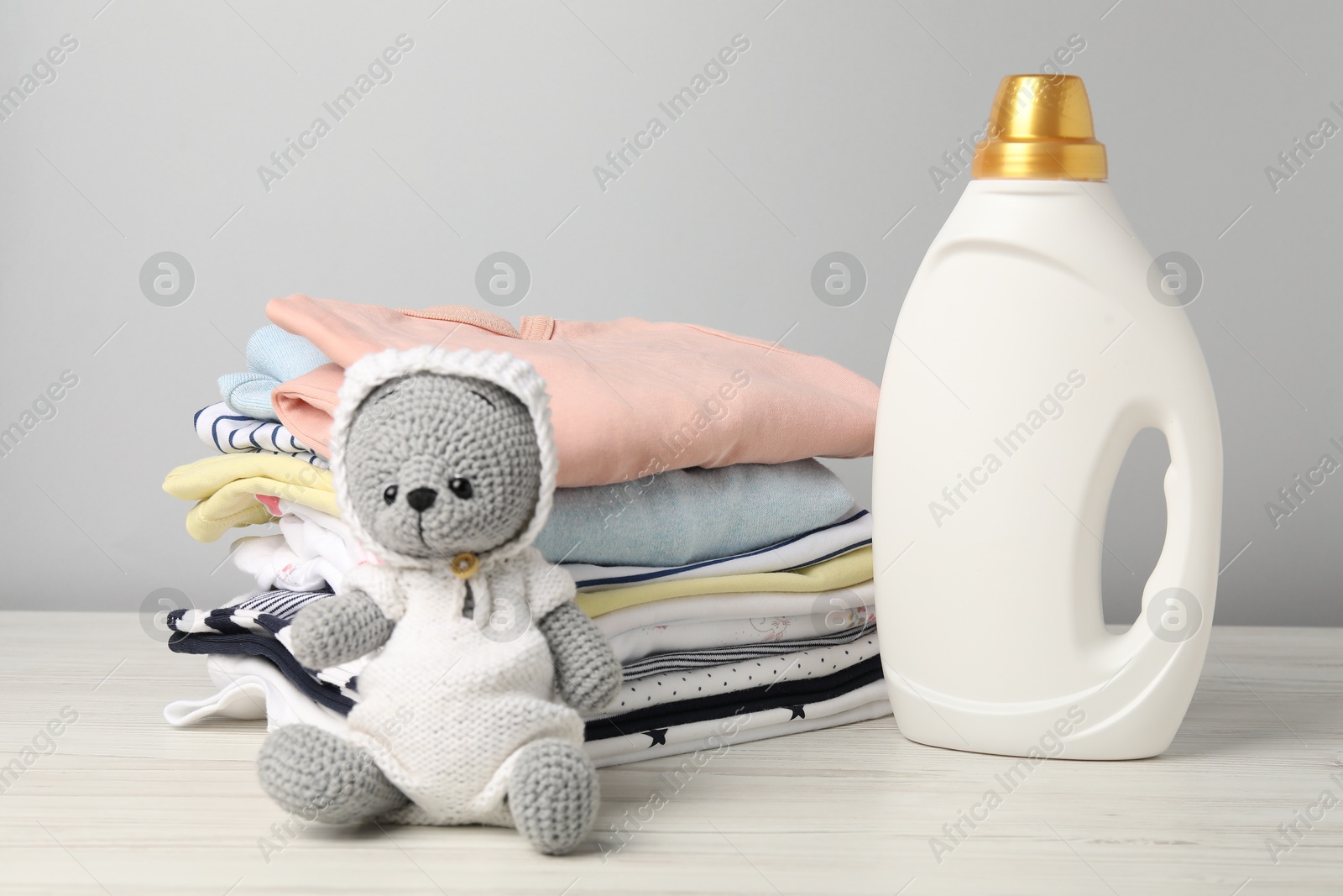 Bottle of fabric softener, baby clothes and toy on light wooden table against grey background Photo of Bottle of fabric softener, baby clothes and toy on light wooden table against grey background