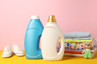 Photo of Bottles of fabric softeners, baby clothes and toy on yellow wooden table against pink background