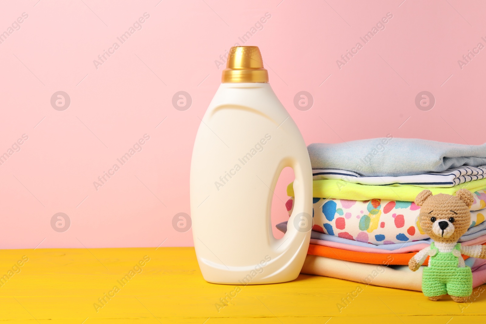 Bottle with fabric softener, stack of baby clothes and toy on yellow wooden table against pink background. Space for text Photo of Bottle with fabric softener, stack of baby clothes and toy on yellow wooden table against pink background. Space for text