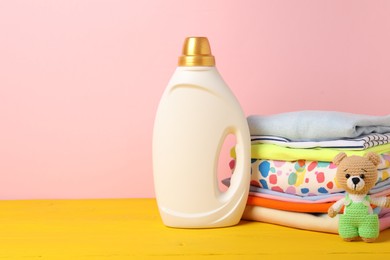 Photo of Bottle with fabric softener, stack of baby clothes and toy on yellow wooden table against pink background. Space for text