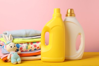 Photo of Bottles of fabric softeners, baby clothes and toy on yellow wooden table against pink background