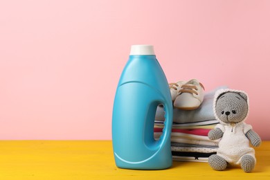 Photo of Bottle with fabric softener, stack of baby clothes and toy on yellow wooden table against pink background. Space for text
