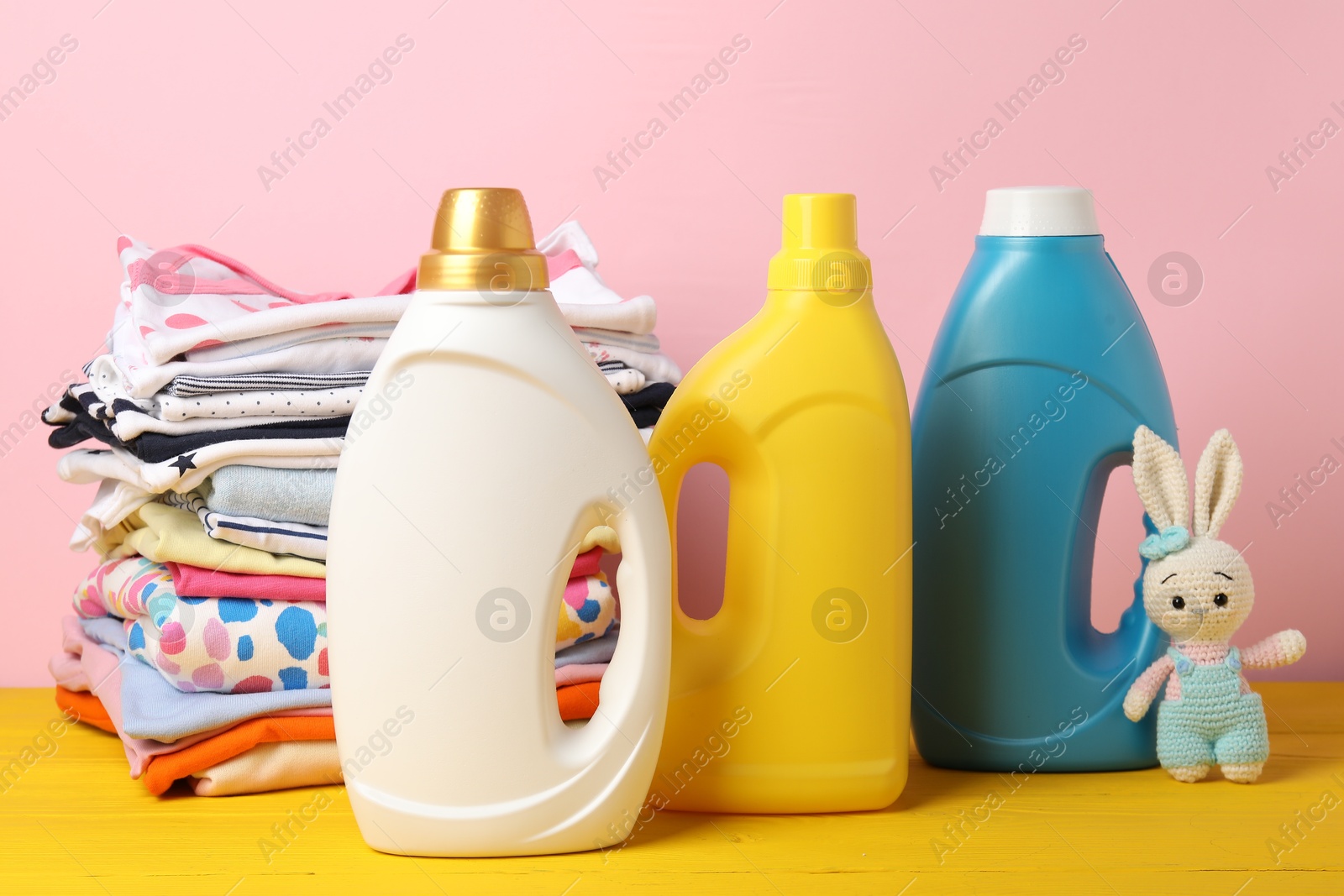 Bottles with fabric softeners, stack of baby clothes and toy on yellow wooden table against pink background Photo of Bottles with fabric softeners, stack of baby clothes and toy on yellow wooden table against pink background