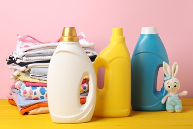 Photo of Bottles with fabric softeners, stack of baby clothes and toy on yellow wooden table against pink background
