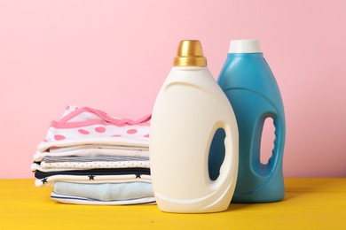 Photo of Bottles with fabric softeners and stack of baby clothes on yellow wooden table against pink background