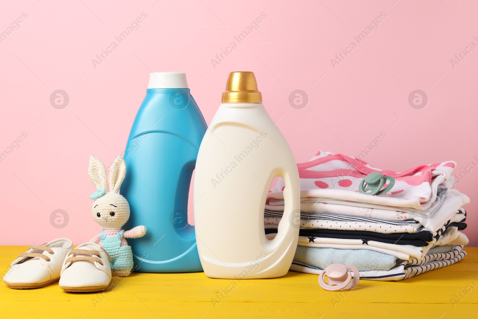 Bottles with fabric softeners, baby clothes, pacifiers and toy on yellow wooden table against pink background Photo of Bottles with fabric softeners, baby clothes, pacifiers and toy on yellow wooden table against pink background