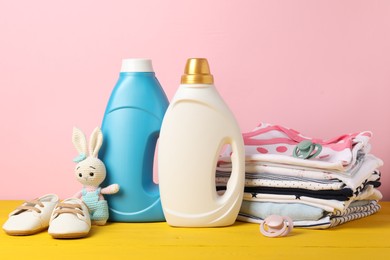 Photo of Bottles with fabric softeners, baby clothes, pacifiers and toy on yellow wooden table against pink background