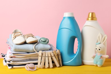 Photo of Bottles with fabric softeners, baby clothes, clothespins and toy on yellow wooden table against pink background