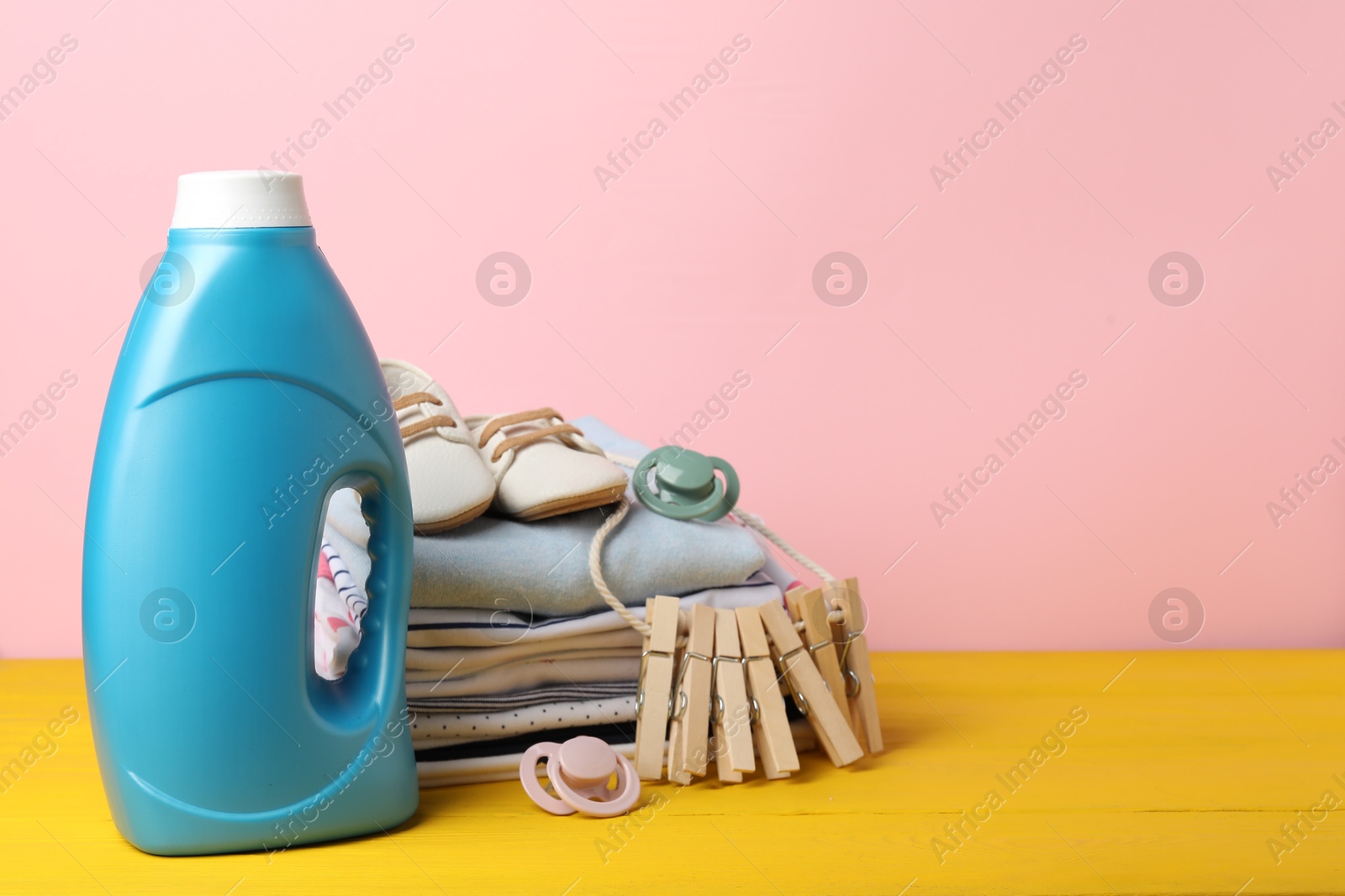 Bottle with fabric softener, baby clothes, pacifiers and clothespins on yellow wooden table against pink background. Space for text Photo of Bottle with fabric softener, baby clothes, pacifiers and clothespins on yellow wooden table against pink background. Space for text