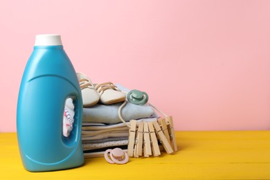 Photo of Bottle with fabric softener, baby clothes, pacifiers and clothespins on yellow wooden table against pink background. Space for text