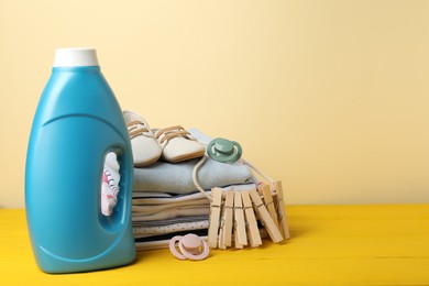 Photo of Bottle with fabric softener, baby clothes, pacifiers and clothespins on yellow wooden table against beige background. Space for text