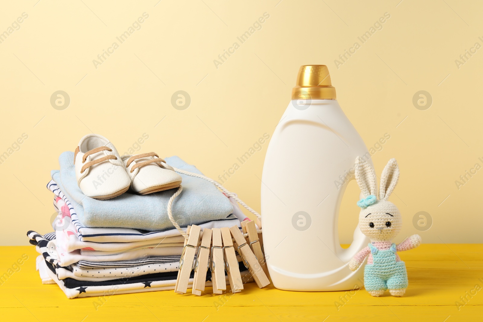 Bottle with fabric softener, baby clothes, clothespins and toy on yellow wooden table against beige background Photo of Bottle with fabric softener, baby clothes, clothespins and toy on yellow wooden table against beige background