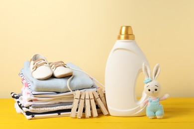 Photo of Bottle with fabric softener, baby clothes, clothespins and toy on yellow wooden table against beige background