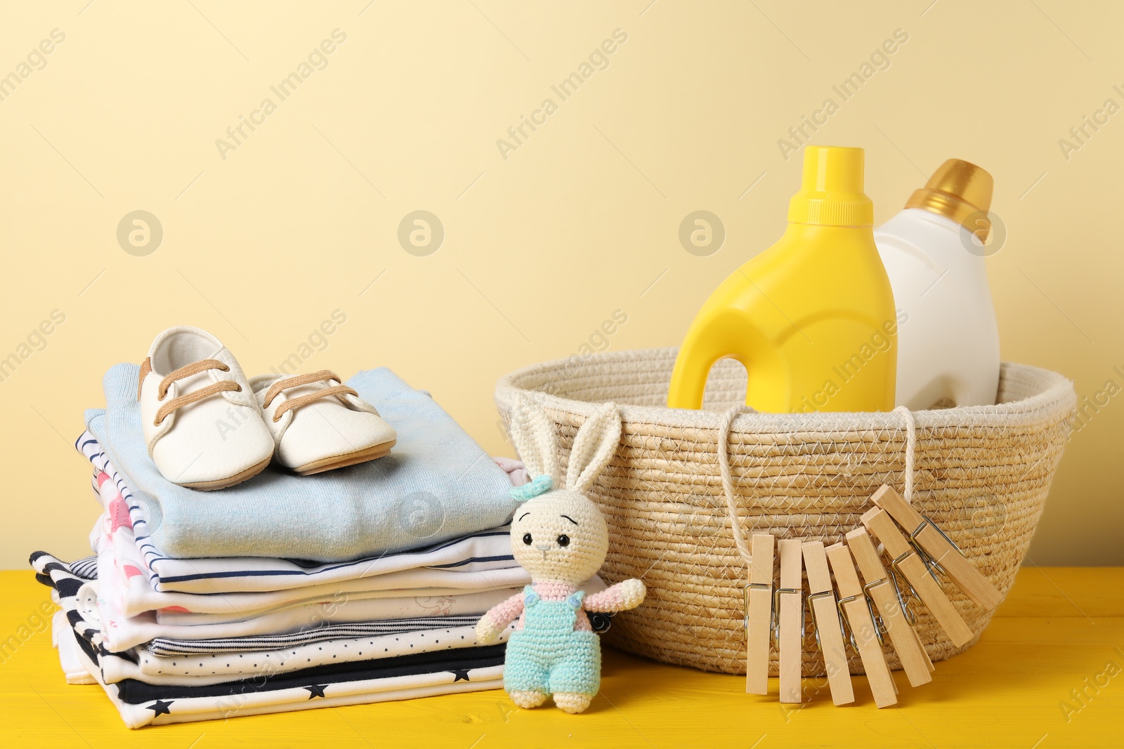 Bottles with fabric softeners, baby clothes, clothespins and toy on yellow wooden table against beige background Photo of Bottles with fabric softeners, baby clothes, clothespins and toy on yellow wooden table against beige background