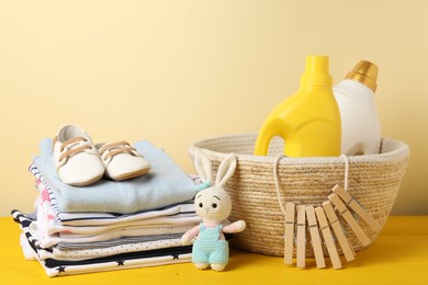 Photo of Bottles with fabric softeners, baby clothes, clothespins and toy on yellow wooden table against beige background