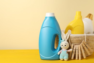 Photo of Bottles with fabric softeners, clothespins and toy on yellow wooden table against beige background. Space for text