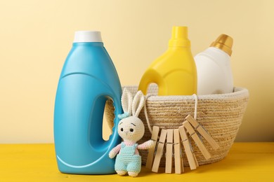 Photo of Bottles with fabric softeners, clothespins and toy on yellow wooden table against beige background