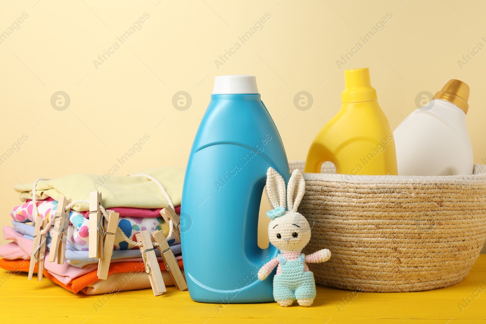 Bottles with fabric softeners, baby clothes, clothespins and toy on yellow wooden table against beige background Photo of Bottles with fabric softeners, baby clothes, clothespins and toy on yellow wooden table against beige background