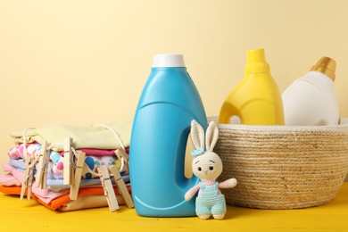 Photo of Bottles with fabric softeners, baby clothes, clothespins and toy on yellow wooden table against beige background