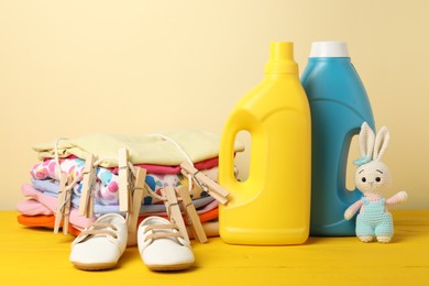 Photo of Bottles with fabric softeners, baby clothes, clothespins and toy on yellow wooden table against beige background