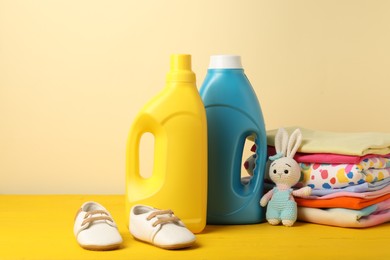 Photo of Bottles with fabric softeners, baby clothes and toy on yellow wooden table against beige background