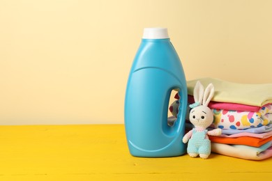 Photo of Bottle with fabric softener, baby clothes and toy on yellow wooden table against beige background. Space for text