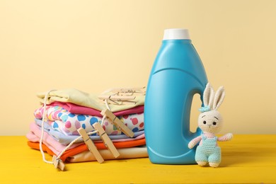 Photo of Bottle with fabric softener, baby clothes, clothespins and toy on yellow wooden table against beige background