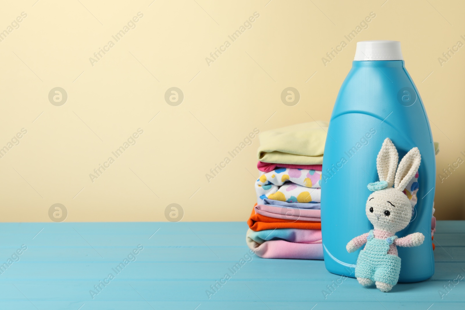 Bottle with fabric softener, baby clothes and toy on light blue wooden table against beige background. Space for text Photo of Bottle with fabric softener, baby clothes and toy on light blue wooden table against beige background. Space for text