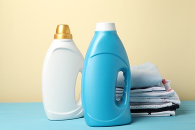 Photo of Bottles with fabric softeners and stack of baby clothes on light blue wooden table against beige background