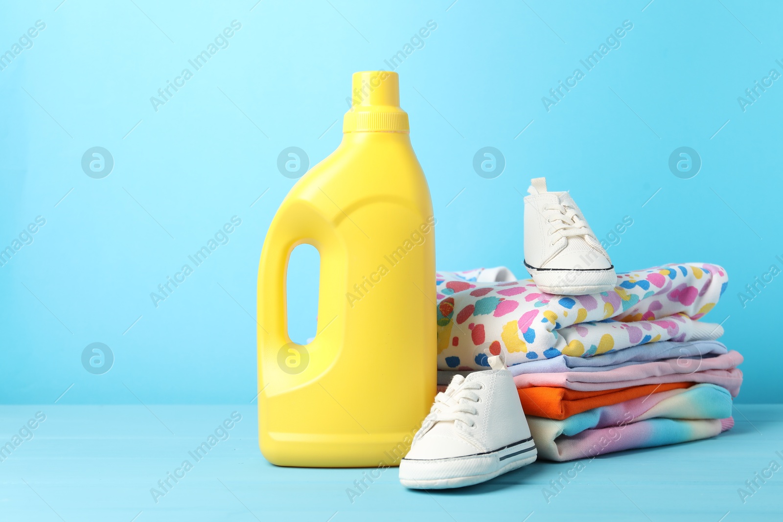 Bottle with fabric softener, baby clothes and shoes on light blue wooden table Photo of Bottle with fabric softener, baby clothes and shoes on light blue wooden table