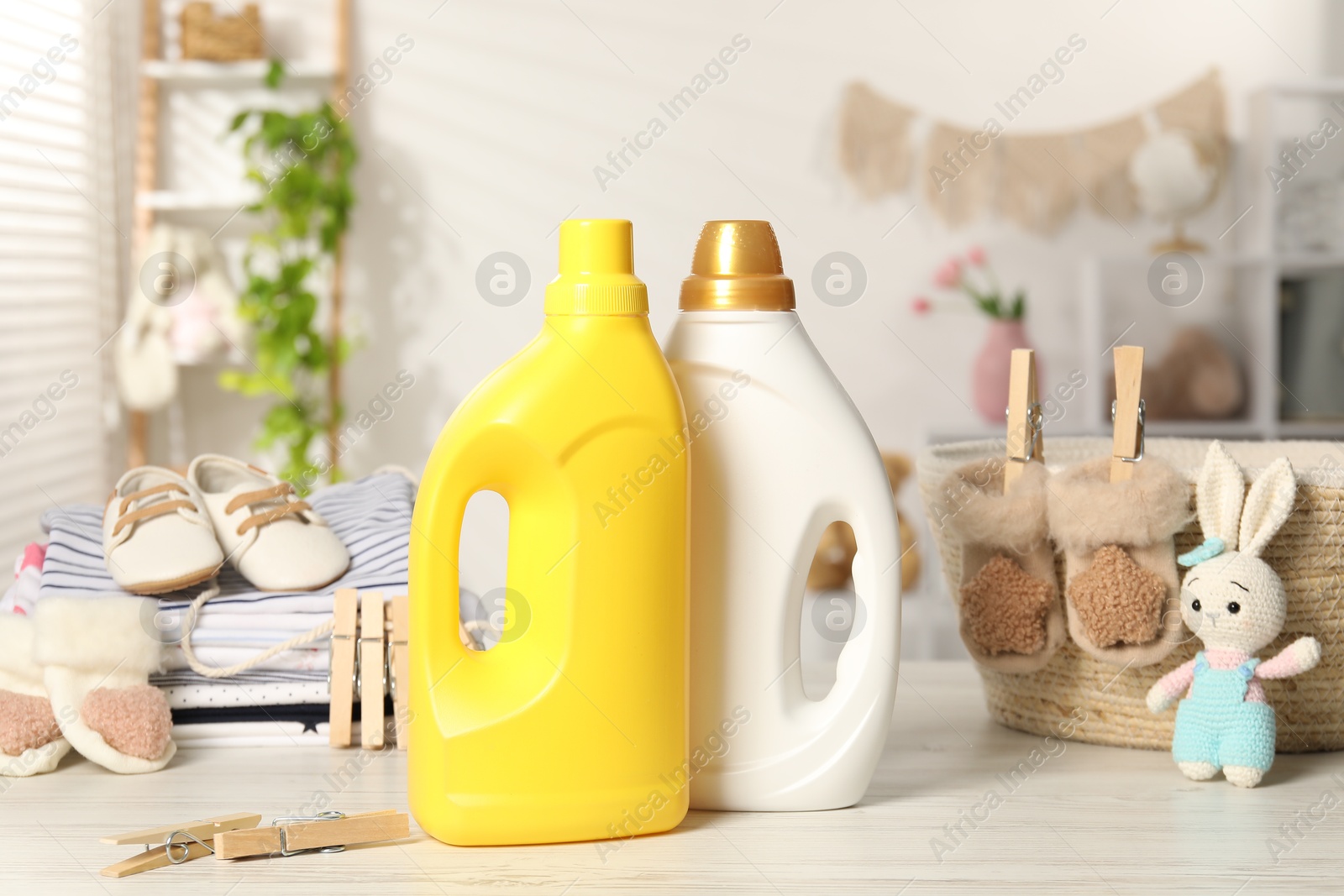 Bottles with fabric softeners, baby clothes and clothespins on light wooden table indoors Photo of Bottles with fabric softeners, baby clothes and clothespins on light wooden table indoors