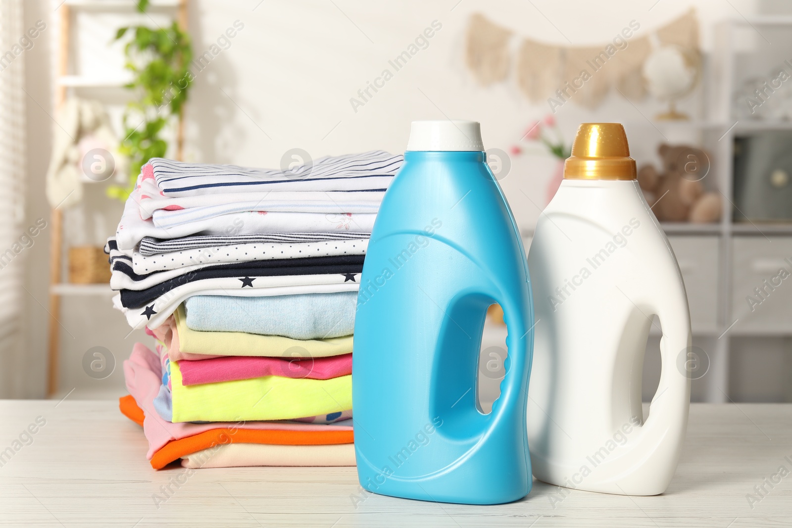 Bottles with fabric softeners and stack of baby clothes on light wooden table indoors Photo of Bottles with fabric softeners and stack of baby clothes on light wooden table indoors