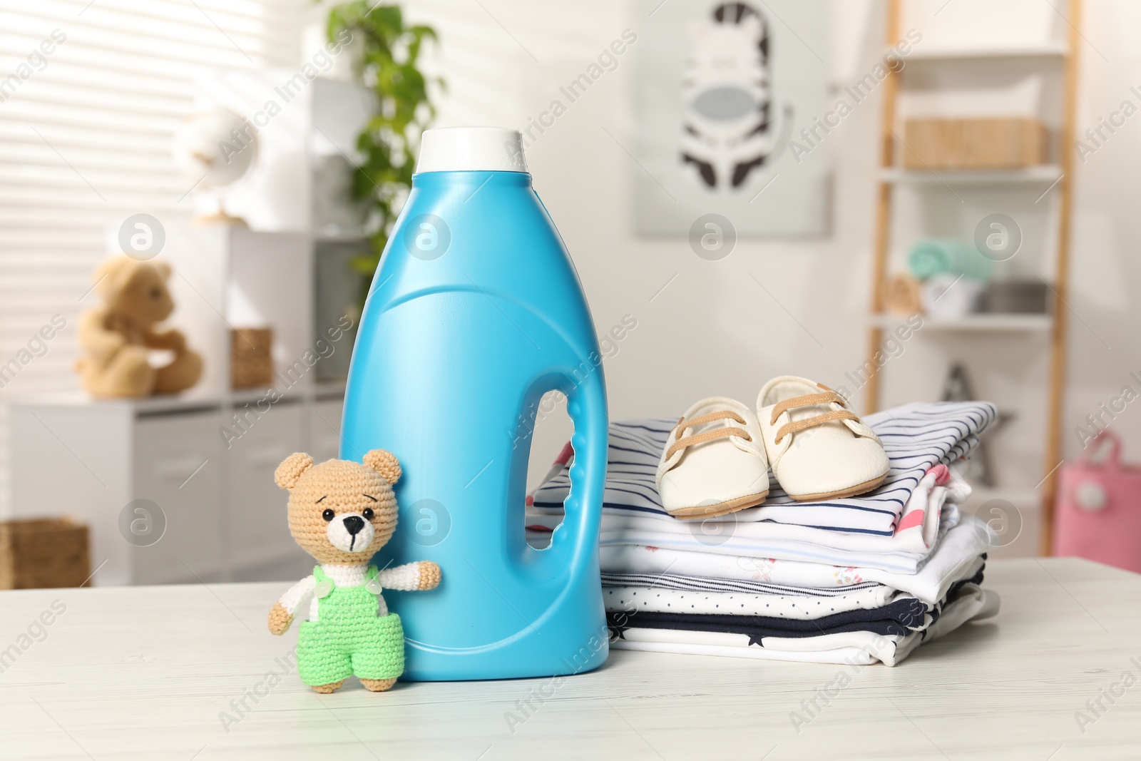 Bottle with fabric softener, stack of baby clothes and toy on light wooden table indoors Photo of Bottle with fabric softener, stack of baby clothes and toy on light wooden table indoors