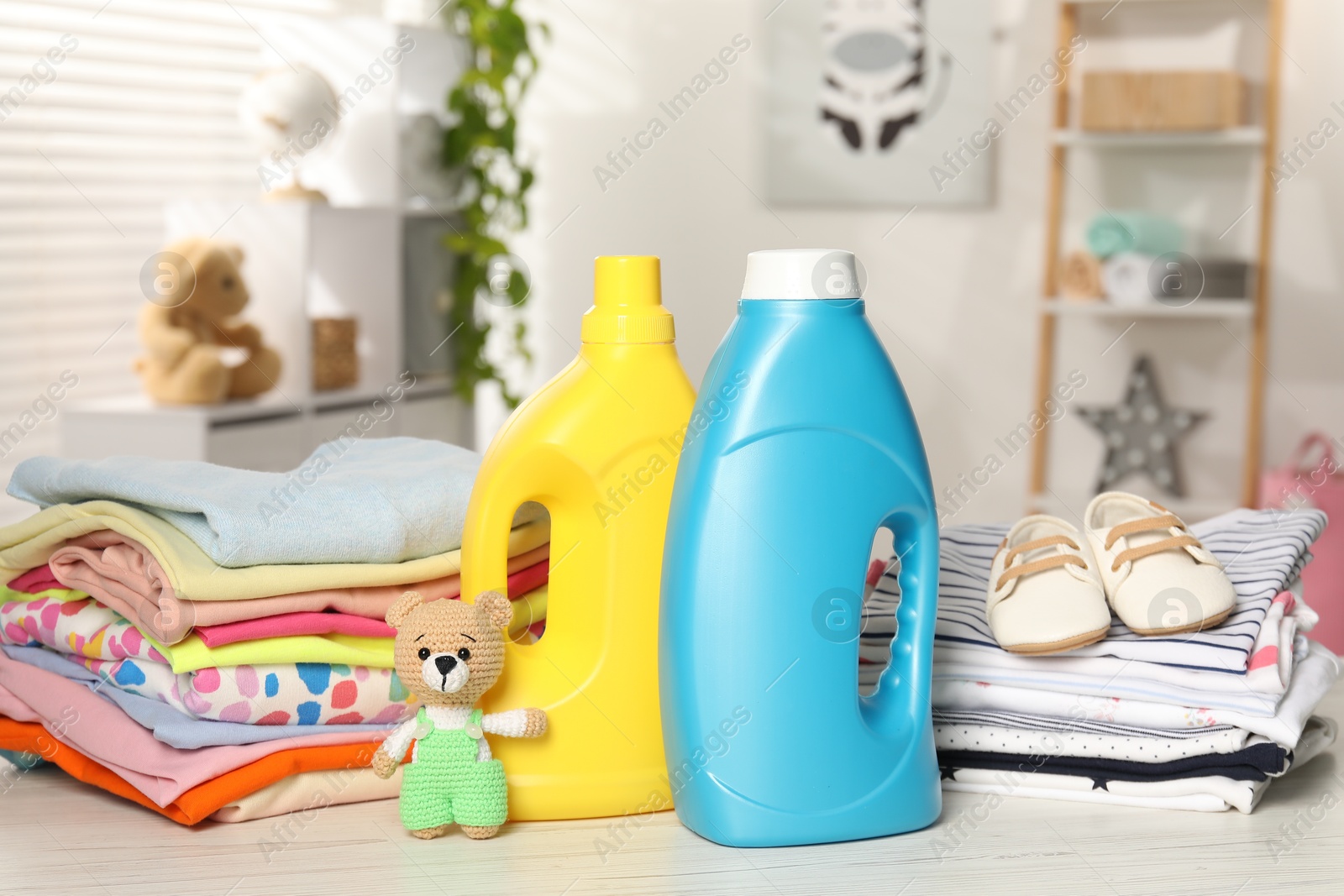 Bottles with fabric softeners, baby clothes and toy on light wooden table indoors Photo of Bottles with fabric softeners, baby clothes and toy on light wooden table indoors