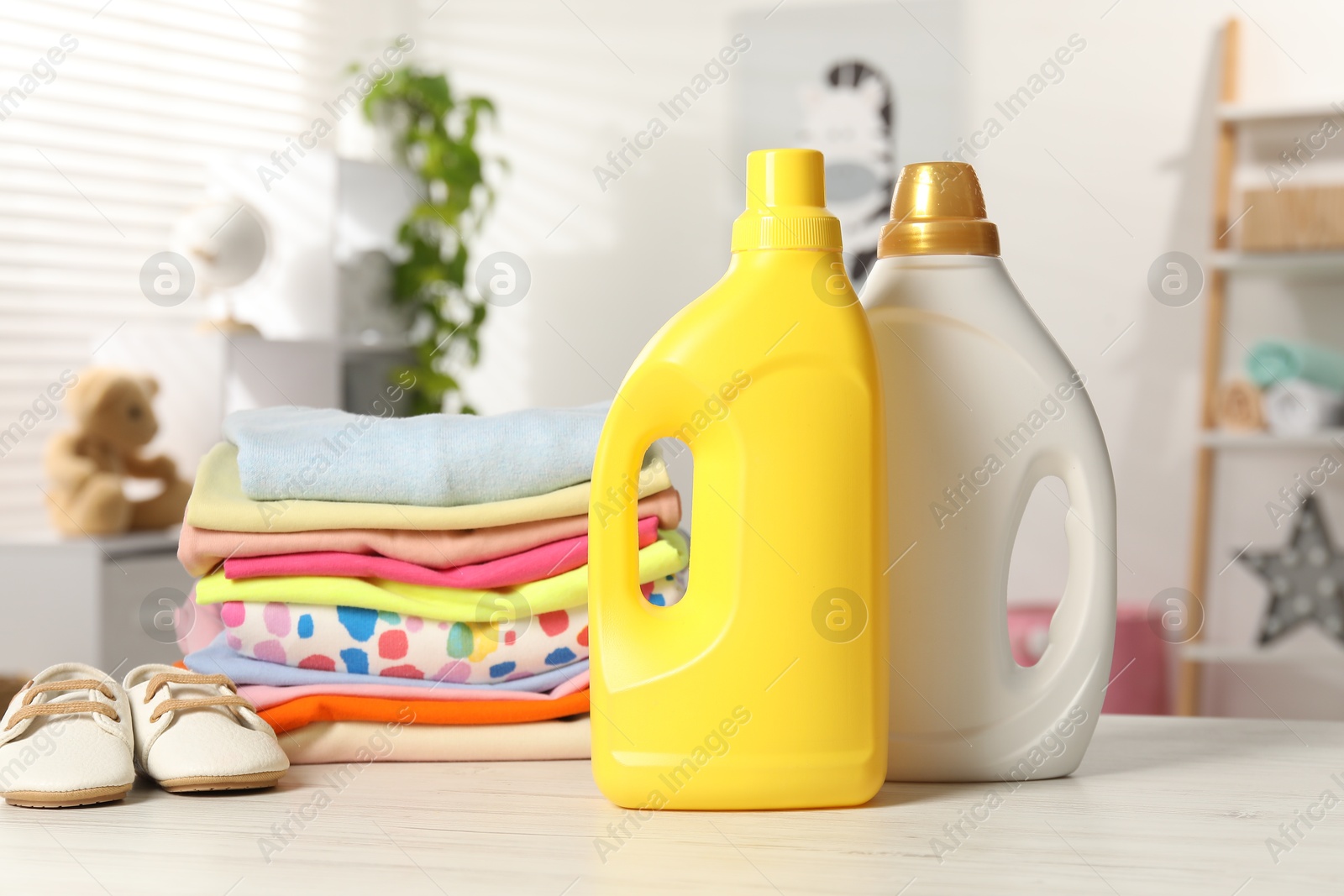 Bottles with fabric softeners and stack of baby clothes on light wooden table indoors Photo of Bottles with fabric softeners and stack of baby clothes on light wooden table indoors