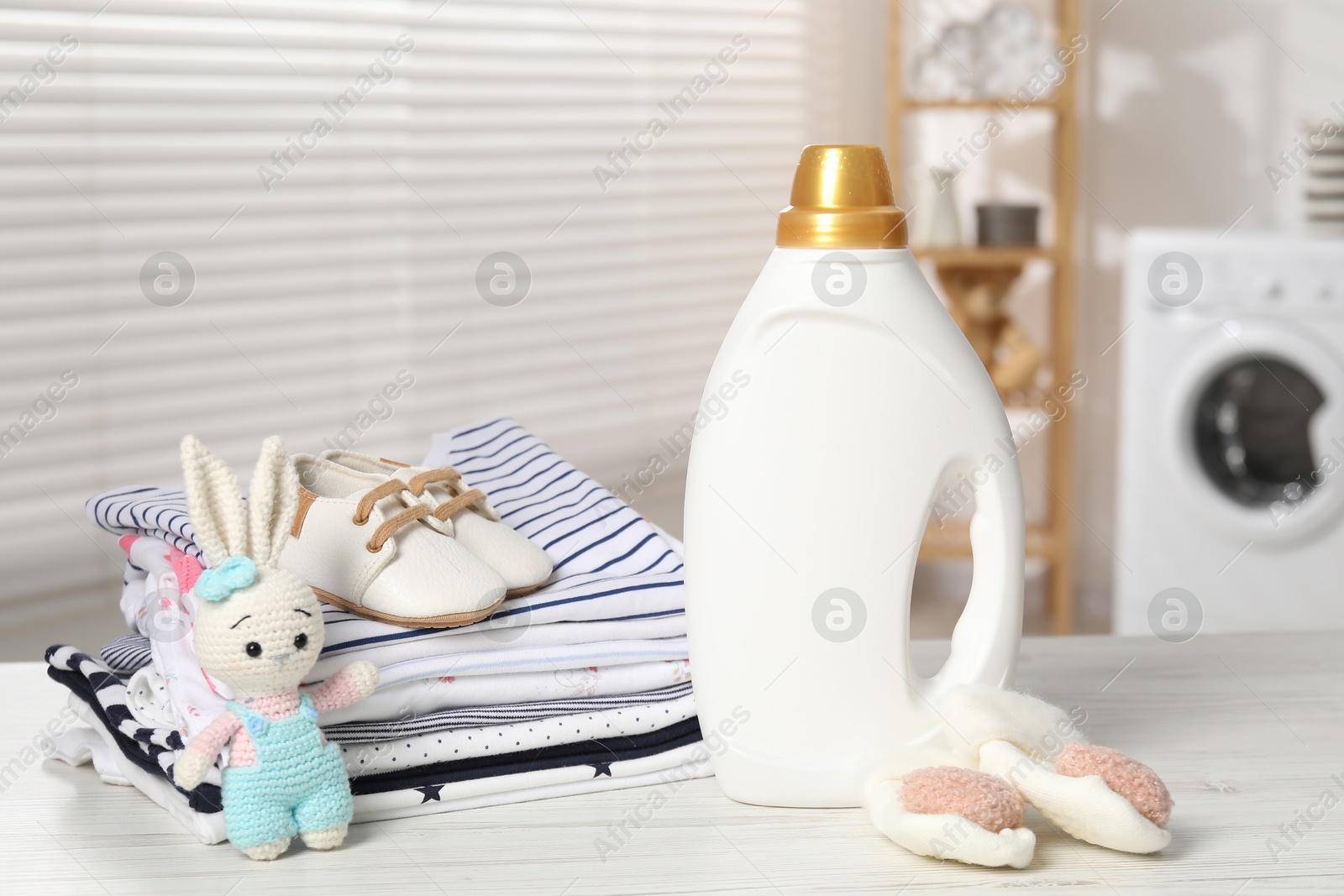 Bottle with fabric softener, baby clothes and toy on light wooden table indoors Photo of Bottle with fabric softener, baby clothes and toy on light wooden table indoors