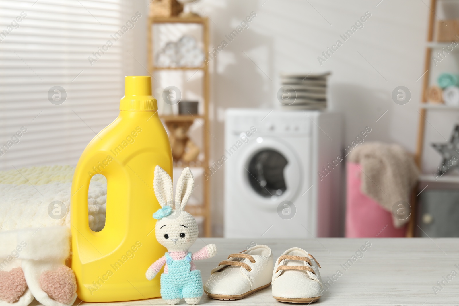 Bottle with fabric softener, baby shoes and toy on light wooden table indoors. Space for text Photo of Bottle with fabric softener, baby shoes and toy on light wooden table indoors. Space for text