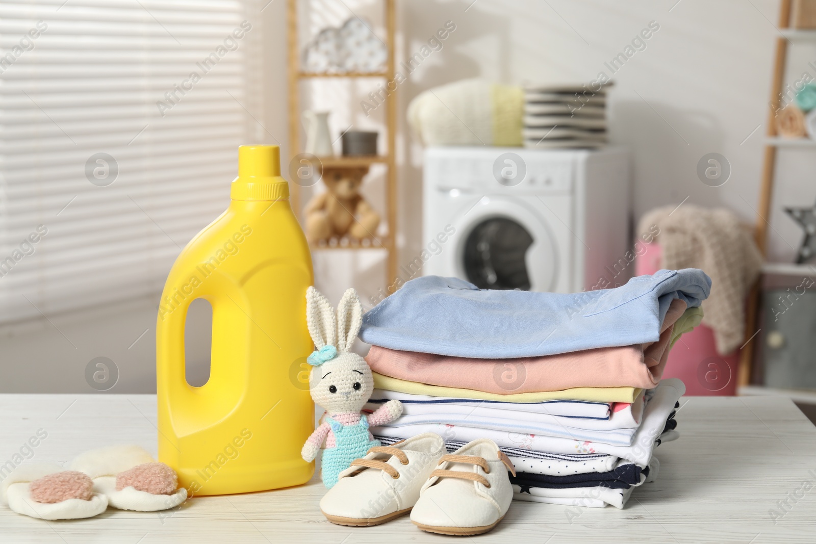 Bottle with fabric softener, baby clothes and toy on light wooden table indoors Photo of Bottle with fabric softener, baby clothes and toy on light wooden table indoors