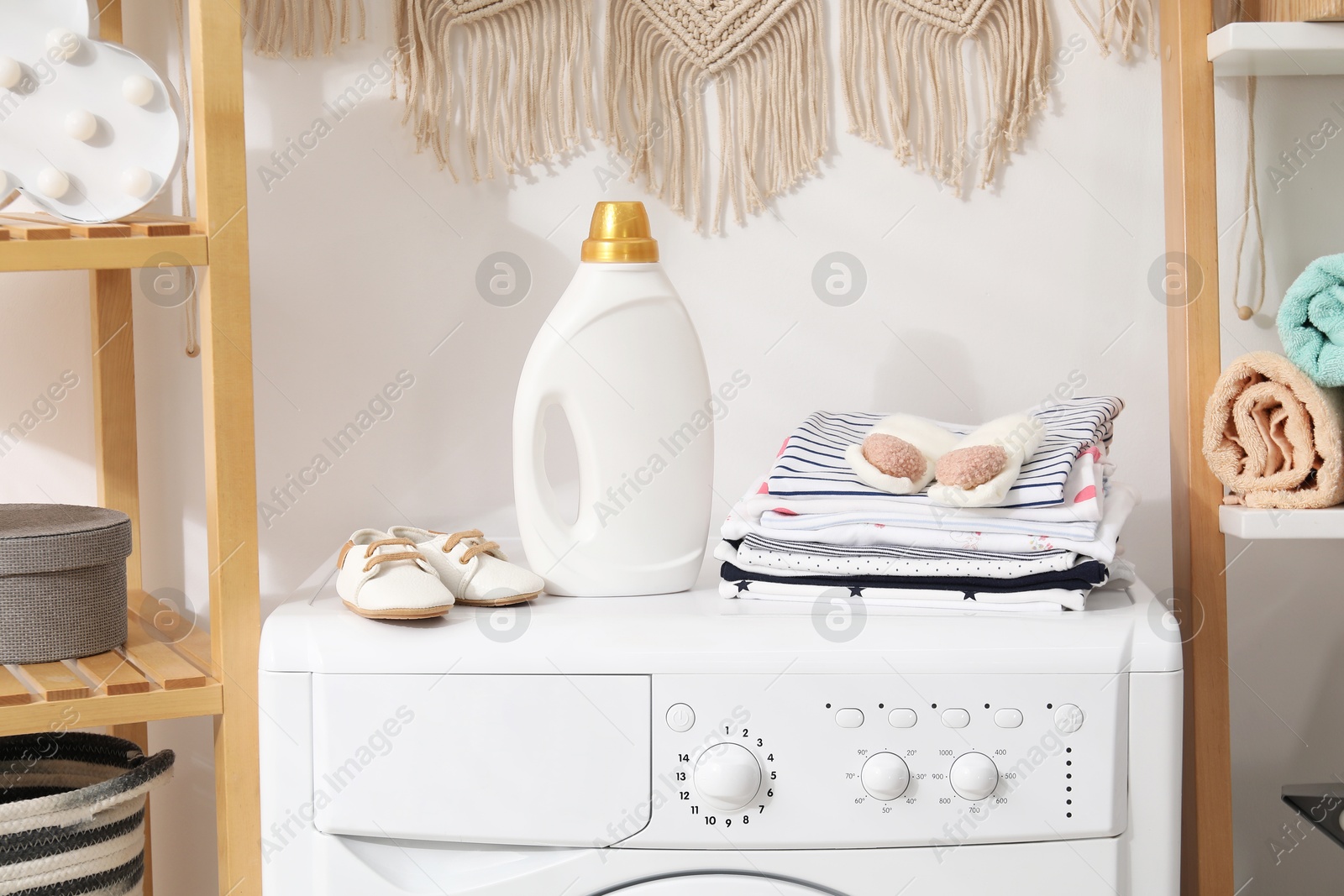 Bottle with fabric softener and baby clothes on washing machine indoors Photo of Bottle with fabric softener and baby clothes on washing machine indoors