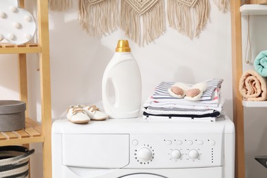 Photo of Bottle with fabric softener and baby clothes on washing machine indoors