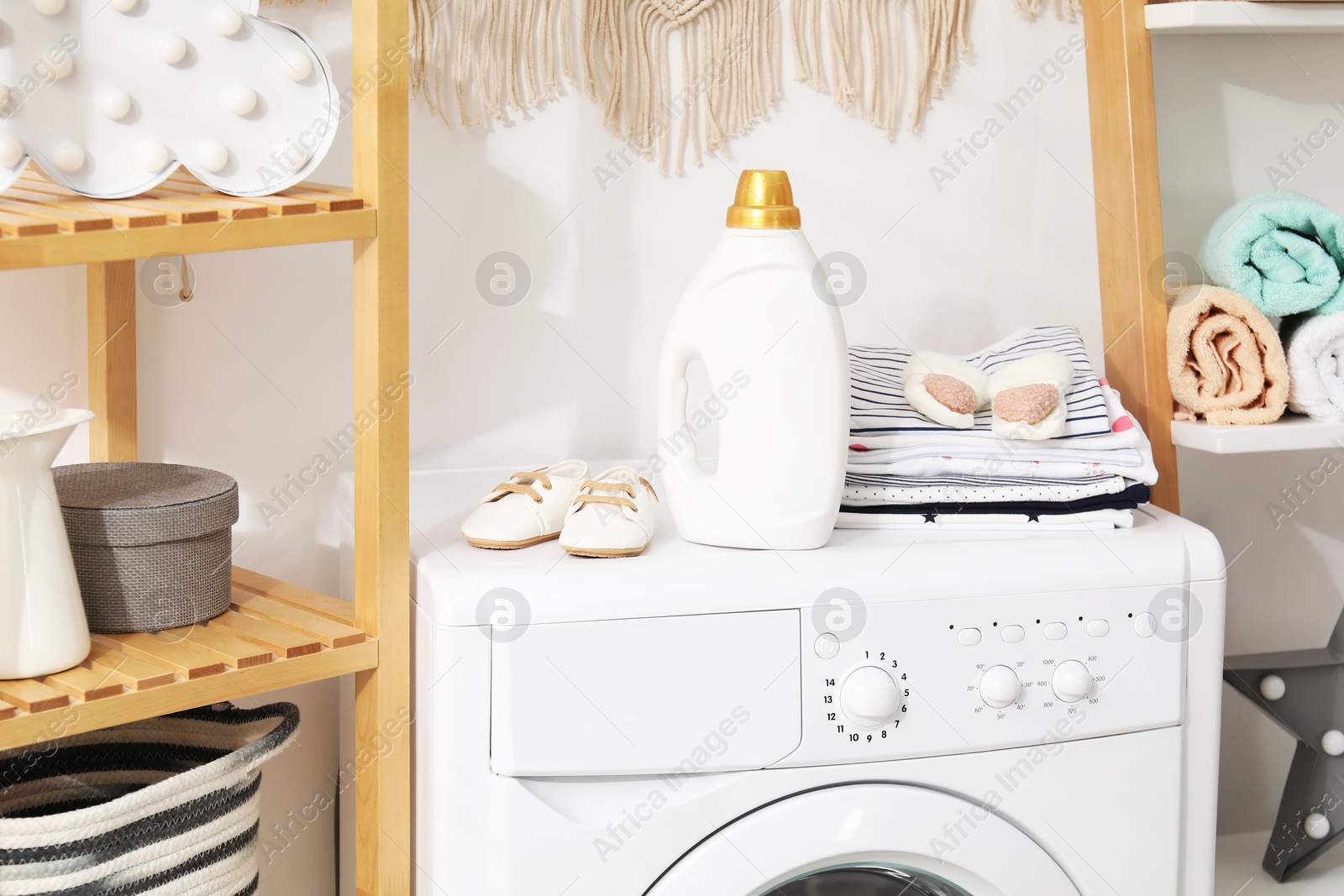 Bottle with fabric softener and baby clothes on washing machine indoors Photo of Bottle with fabric softener and baby clothes on washing machine indoors