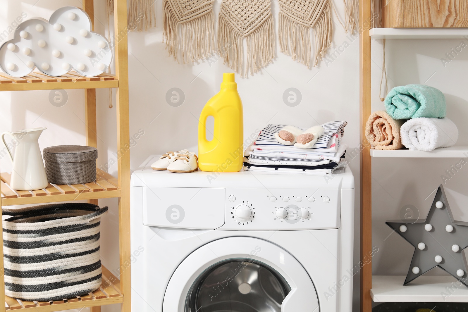 Bottle with fabric softener and baby clothes on washing machine indoors Photo of Bottle with fabric softener and baby clothes on washing machine indoors