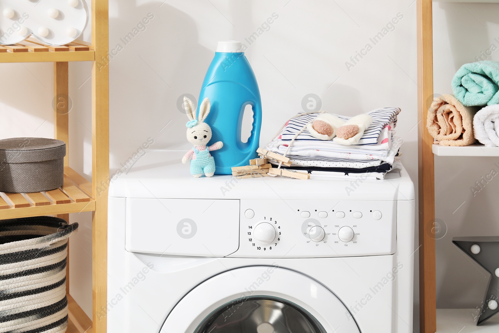 Bottle with fabric softener, baby clothes, clothespins and toy on washing machine indoors Photo of Bottle with fabric softener, baby clothes, clothespins and toy on washing machine indoors