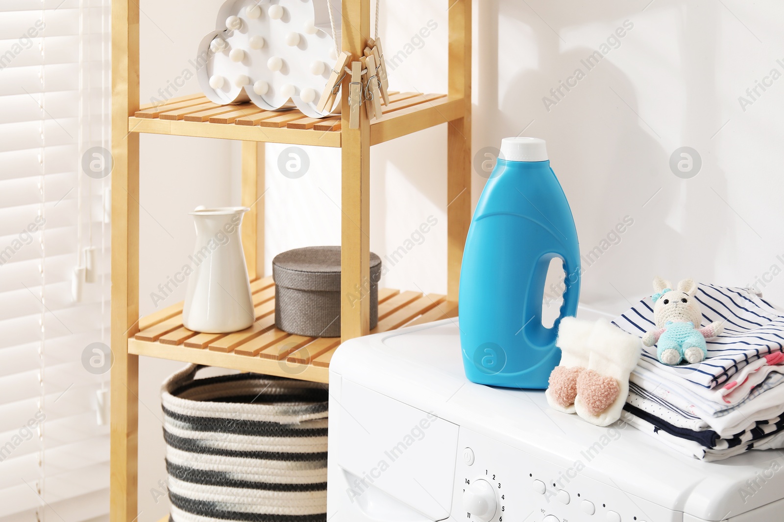 Bottle with fabric softener, baby clothes and toy on washing machine indoors Photo of Bottle with fabric softener, baby clothes and toy on washing machine indoors