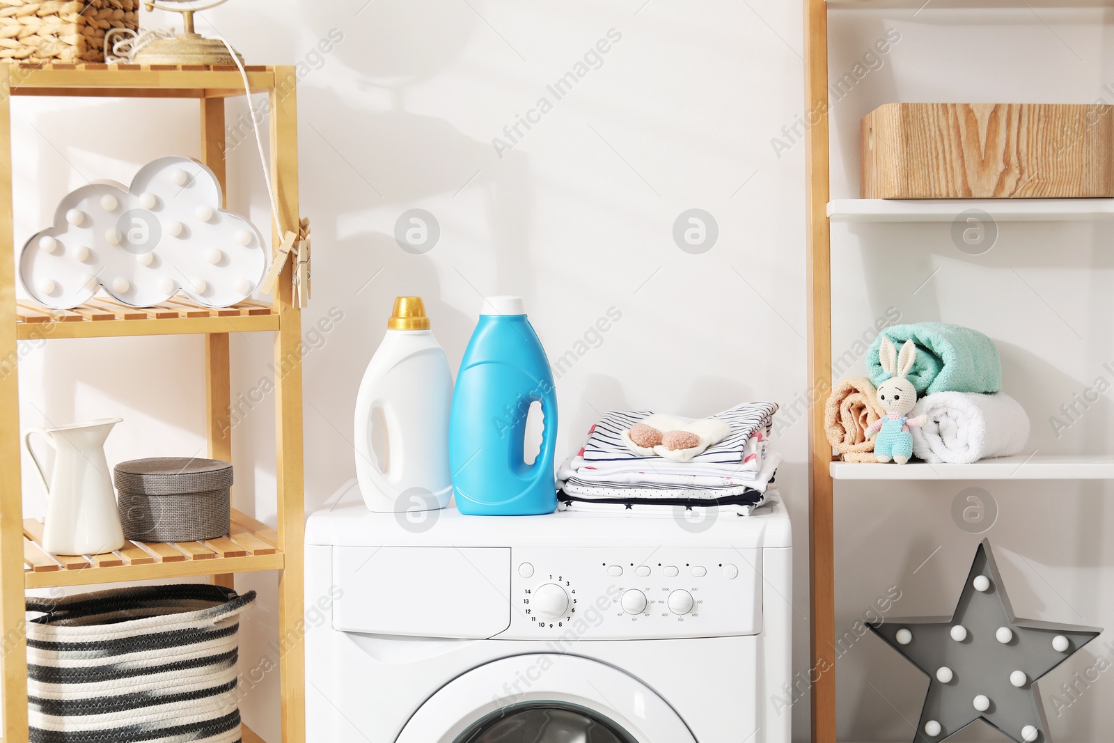 Bottles with fabric softeners and baby clothes on washing machine indoors Photo of Bottles with fabric softeners and baby clothes on washing machine indoors