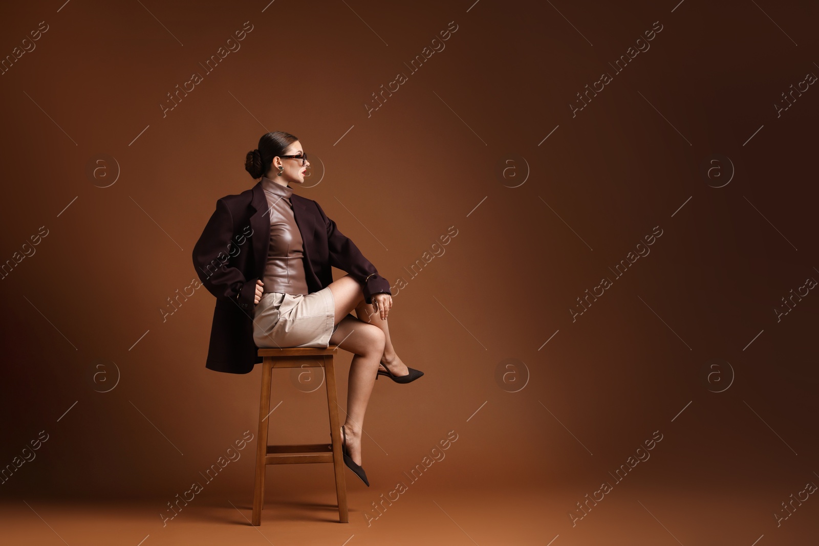 Young woman in stylish clothes on chair against brown background, space for text Photo of Young woman in stylish clothes on chair against brown background, space for text